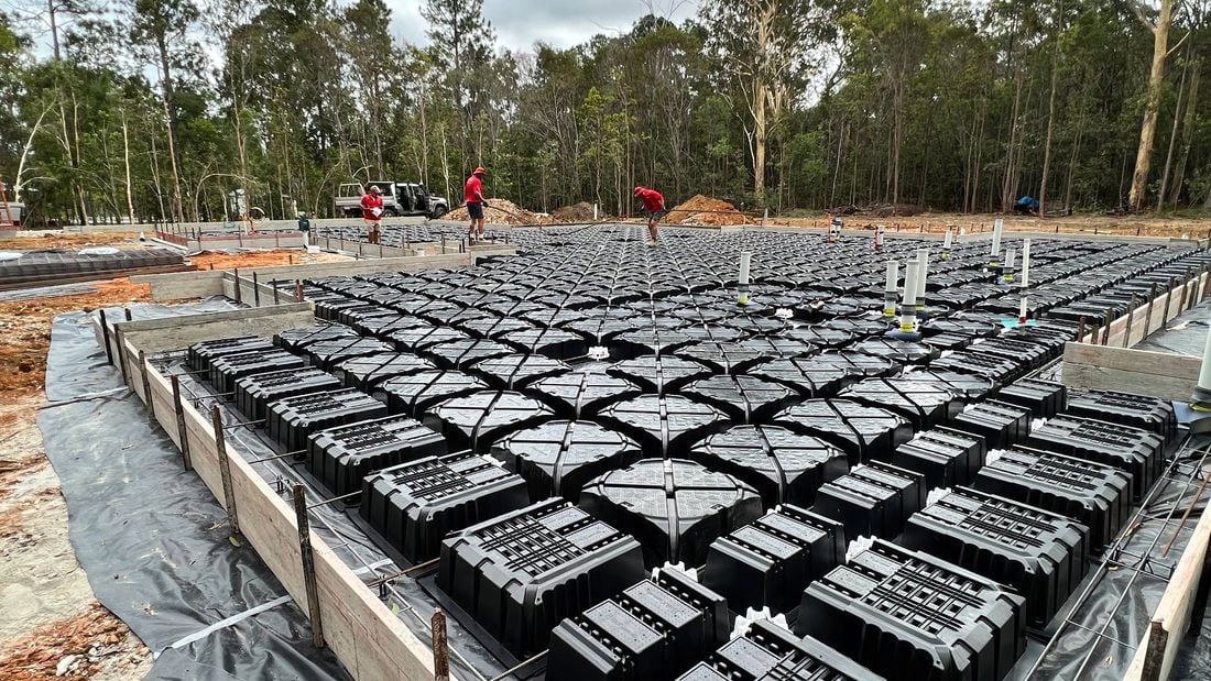 A construction site with a large grid of Biax pods inside timber framework, three tradespeople in red shirts walk across the setup, with bushland in the background