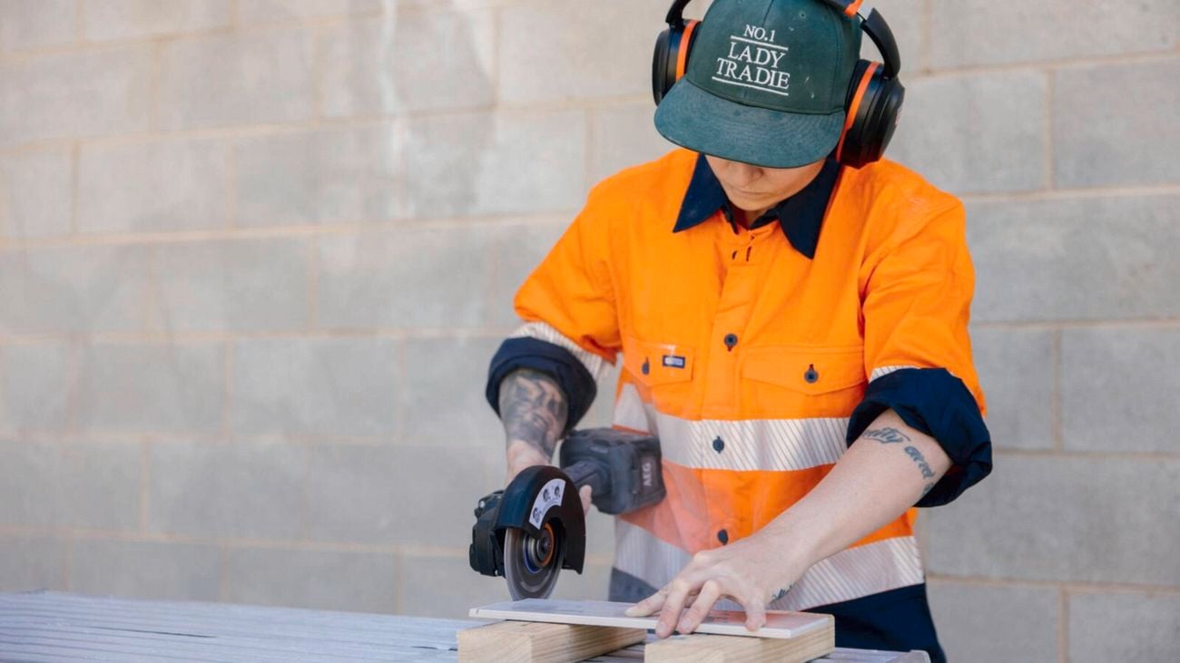 A tradesperson in high-visibility workwear, a cap, and earmuffs uses an angle grinder to cut a tile.