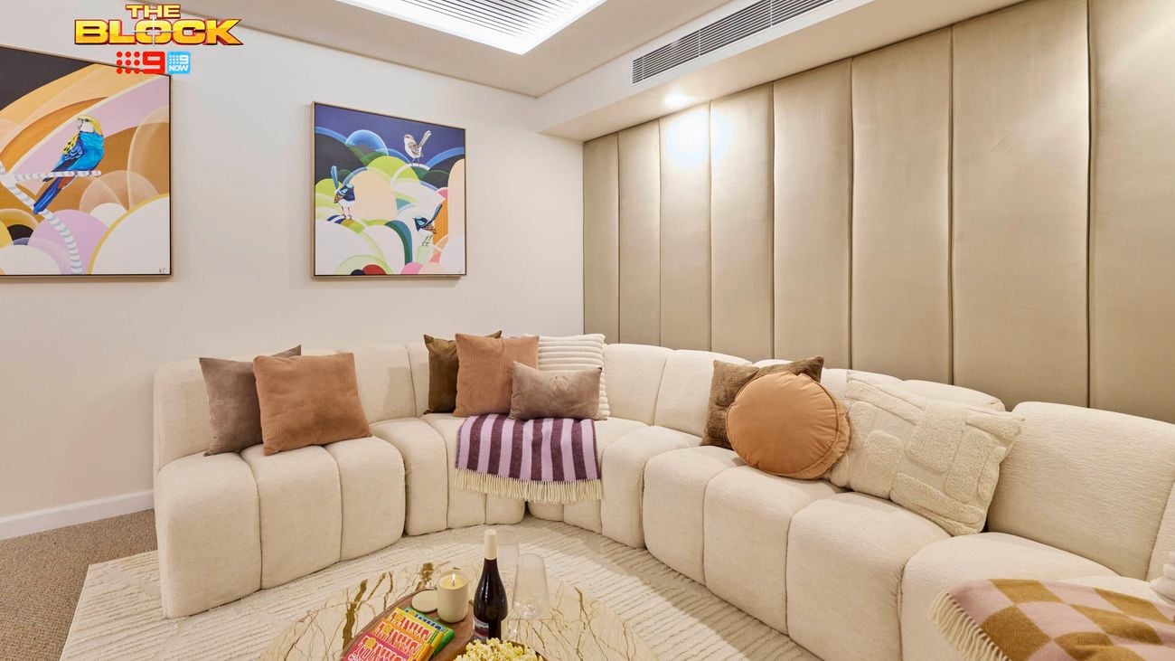 A cozy living room with a beige sectional sofa decorated with assorted throw pillows and blankets. A round coffee table in the center holds popcorn, books, and bottles. The walls feature two colorful abstract paintings, and the ceiling has recessed lighting, contributing to the room’s modern and inviting atmosphere.