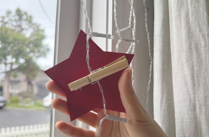 A hand using a wooden peg to attach a red-painted craft star to a string of fairy lights in front of a window.] 