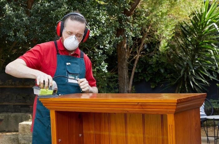 A man wearing safety equipment starts sanding a bookcase with 120-grit sander.