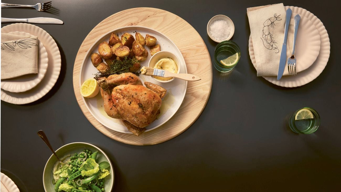 A charcoal chicken with roasted potatoes, a lemon half, and a butter brush on a wooden platter, alongside a bowl of green peas.