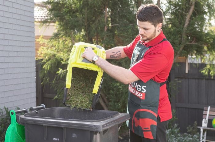 Person emptying lawn mower compartment into green bin.