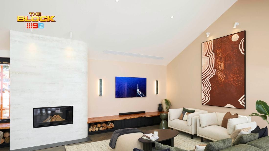 A modern living room with a high ceiling, featuring a fireplace on the left with firewood stored beneath. The space includes a green sofa, white armchairs, and a small round coffee table with books. Two large pieces of wall art are displayed—one above the fireplace and another on the right wall. 