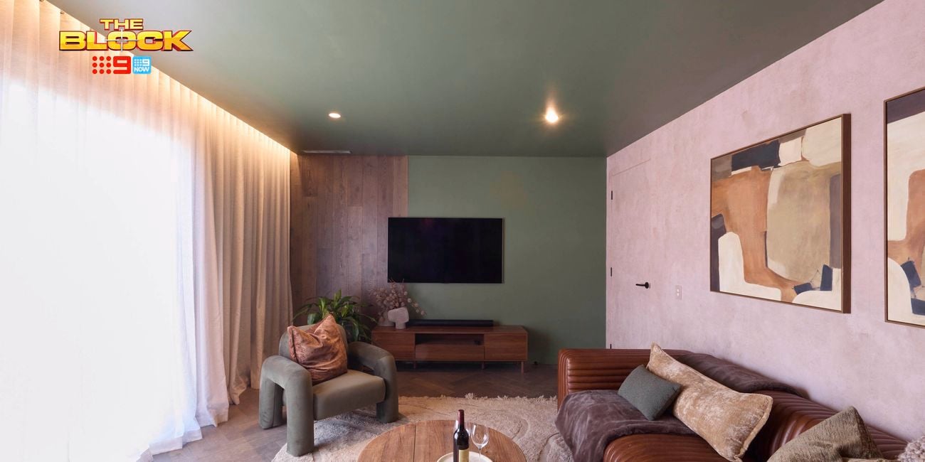 A modern living room with a green ceiling and light-colored walls, featuring a large window with sheer white curtains. The space includes a brown leather sofa with cushions, a green armchair, and two wooden coffee tables—one holding a tray with a bottle of wine and two glasses. A flat-screen TV is mounted above a wooden media console, and two abstract paintings hang above the sofa. A textured rug covers part of the floor.