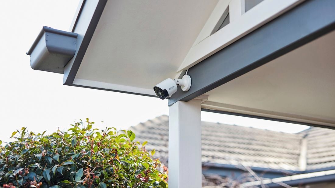 White security camera installed under a house roof eave above an outdoor area, with shrubs and neighbouring rooftops visible in the background.
