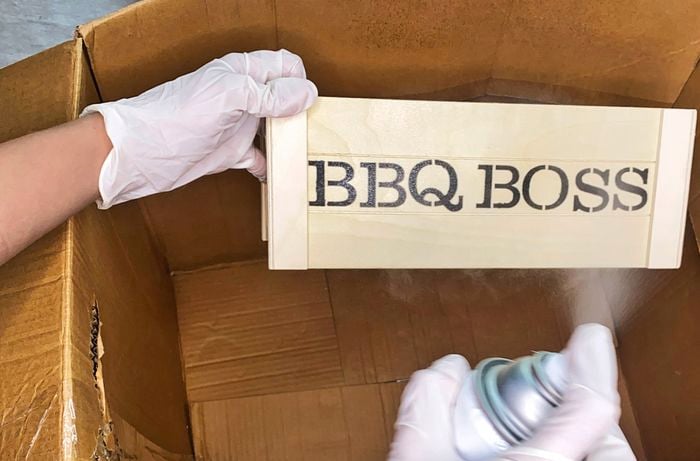 In a cardboard box, a gloved hand sprays clear sealer on a stencilled small timber crate.