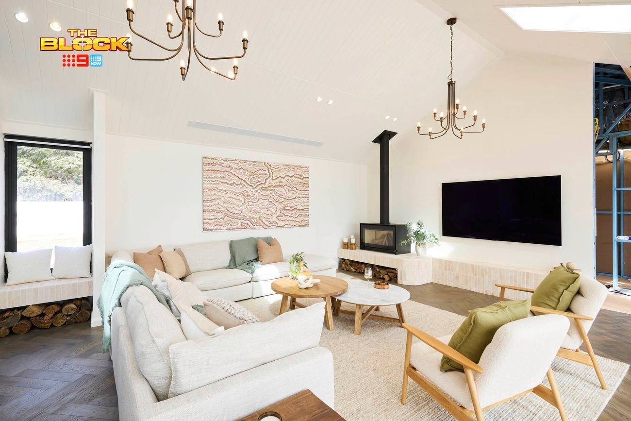 A modern living room with high ceilings, featuring two chandeliers, a wall-mounted flat-screen TV, and a fireplace with logs stored below. The space includes a light-colored sectional sofa with cushions and throws, two armchairs with green cushions, and two round wooden coffee tables. Abstract artwork hangs above the sofa, and large windows bring in natural light. Decorative plants are placed throughout. 