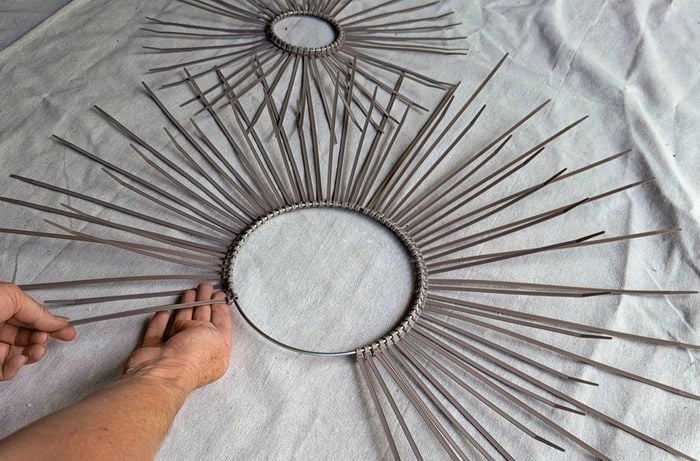 Two circular metal rings covered with evenly spaced cable ties arranged like sun rays, as a hand adds another cable tie to complete the radial pattern.