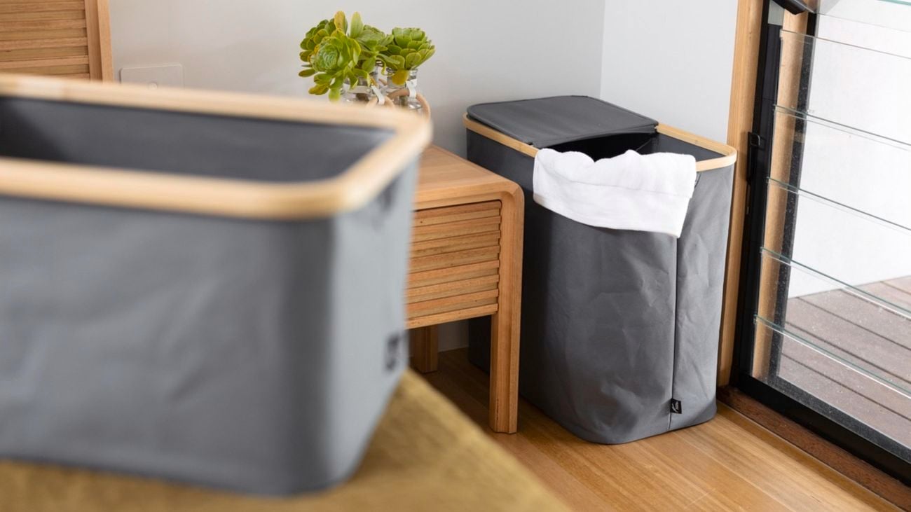 A bedroom featuring a grey Hill bamboo laundry hamper next to a wooden bedside table and a window. Another hamper is out of focus on the bed