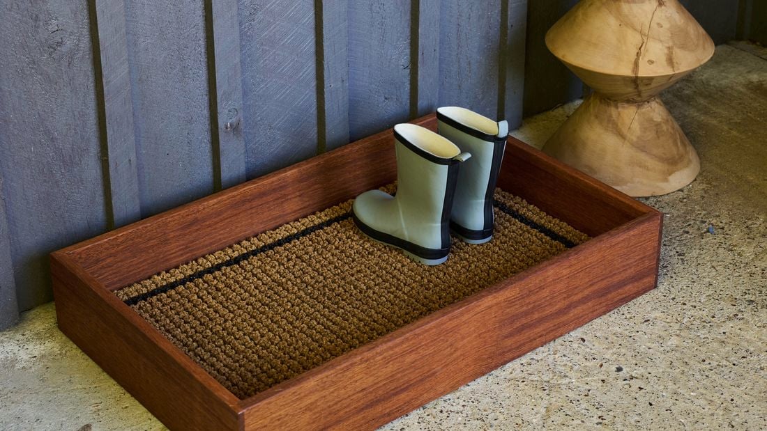 Brown wooden tray with gumboots on it, on the ground, with a fence behind it.