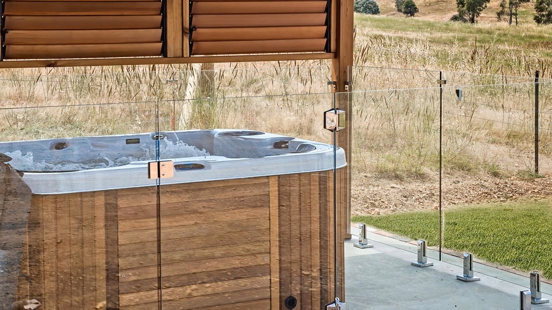 An above ground spa fenced off with glass under a pergola on a back lawn