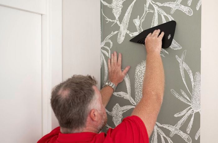 A person in a red T shirt is using a wallpaper smoother to attach the first piece of wallpaper on the wall