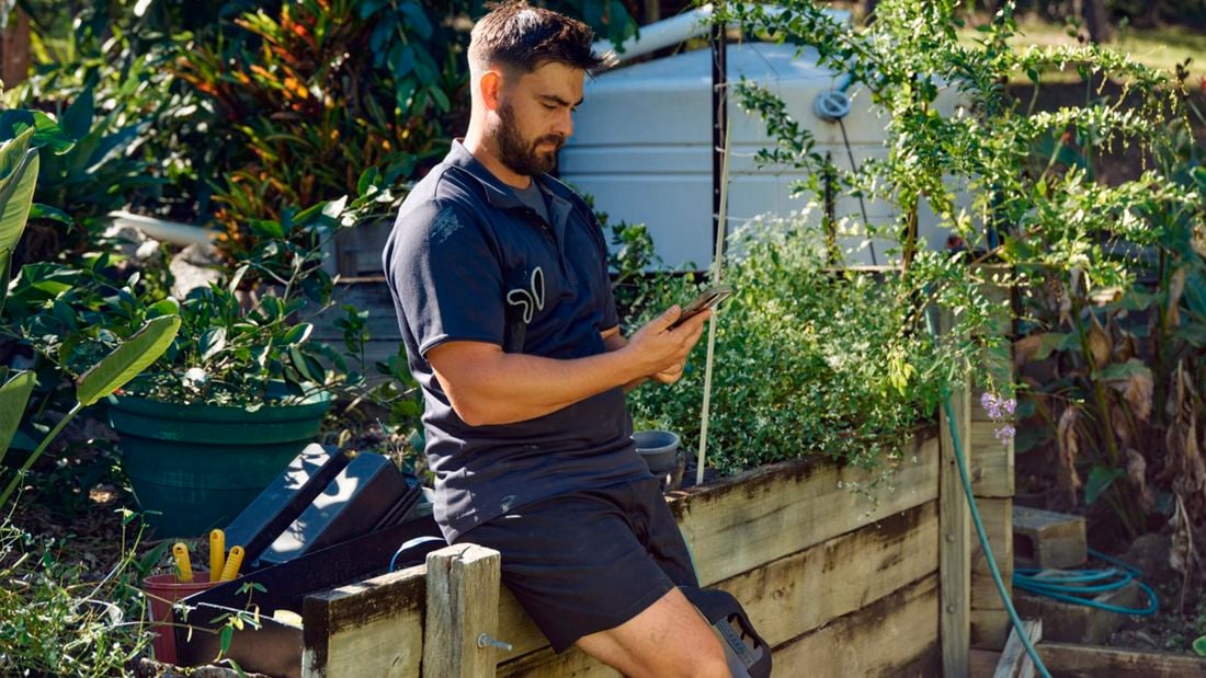Alt tag: A tradie A tradie wearing a dark shirt and shorts sits on a wooden garden bed looking at his mobile phone, surrounded by plants and gardening tools