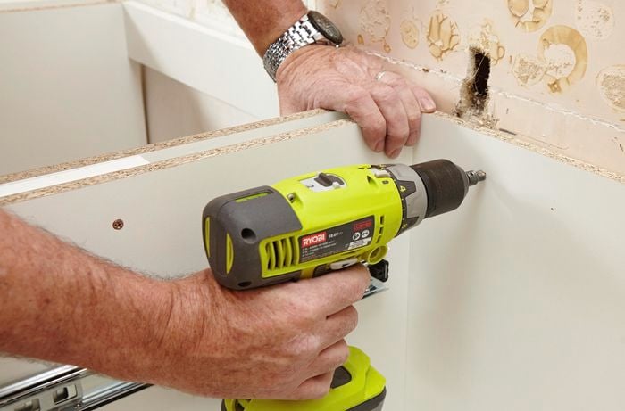 A kitchen cabinet being drilled into place against a bare wall with a power drill