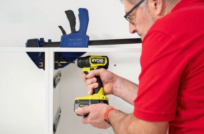 A person is using a power drill to join two cabinet frames together while clamps hold them in place for alignment.