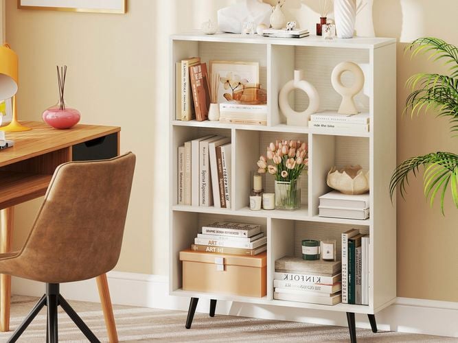 A white bookcase with books, candles, vases with flowers in them in it, and a chair and desk next to it in the room.