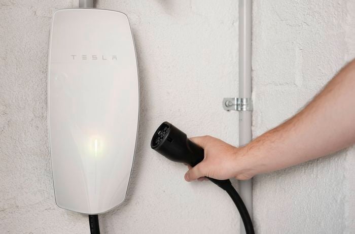 A person is holding a charging cable beside a wall-mounted Tesla EV charger, evaluating the ideal spot for EV charger installation.  