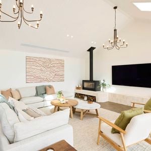 A modern living room with high ceilings, featuring two chandeliers, a wall-mounted flat-screen TV, and a fireplace with logs stored below. The space includes a light-colored sectional sofa with cushions and throws, two armchairs with green cushions, and two round wooden coffee tables. Abstract artwork hangs above the sofa, and large windows bring in natural light. Decorative plants are placed throughout. 