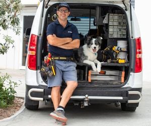 A tradesperson leaning against the back of an open van with tools and equipment visible inside, alongside a dog sitting inside the van.