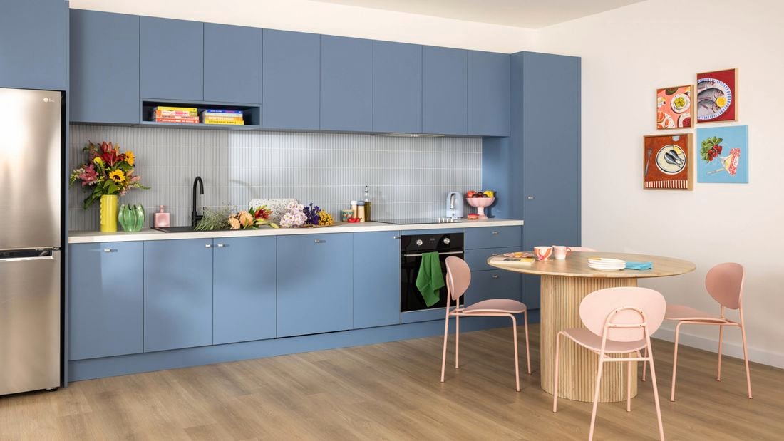 Modern kitchen with blue cabinets, light wooden floor, stainless steel fridge, countertop with flowers, fruits, and appliances, round wooden dining table with four pink chairs, and four colorful food-themed framed pictures on the wall.