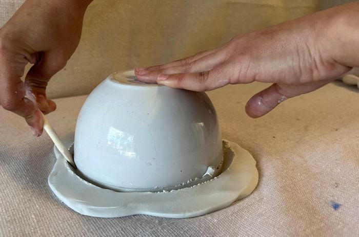 A hand uses a wooden craft tool to cut around an upturned bowl on a clay disc.