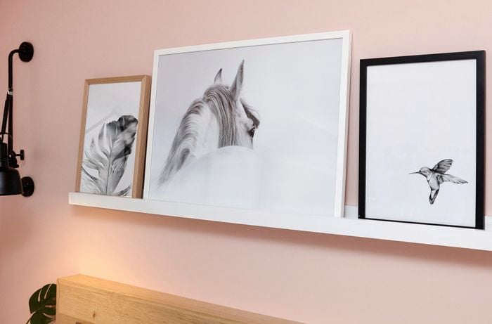 A floating white shelf with framed pictures above a bedhead against a pink wall