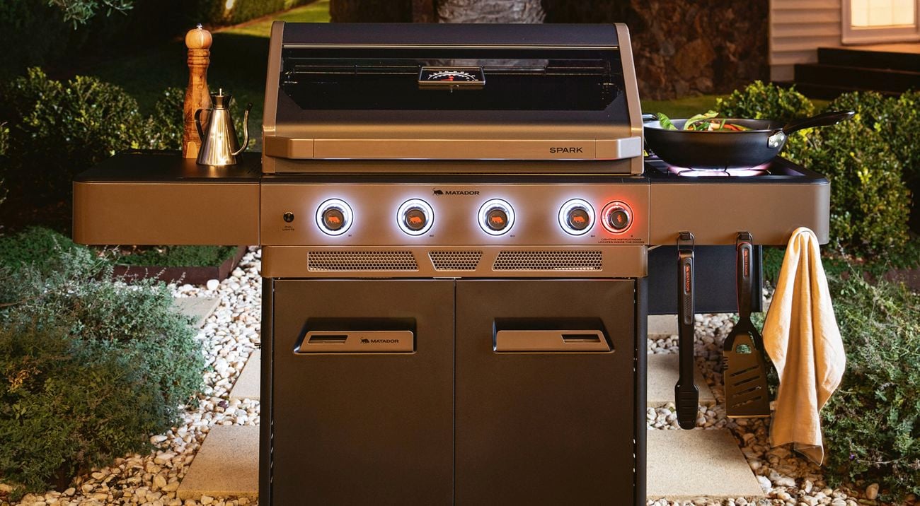 Outdoor barbecue grill with illuminated control knobs, side shelves holding utensils and condiments, set on a stone patio surrounded by greenery and a large palm tree in the background.