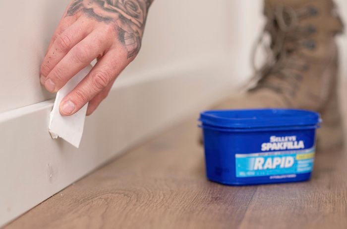 A person is using a scraper to apply gap filler paste to a gap in the skirting board, with a tub of Selleys Spakfilla Rapid nearby. 