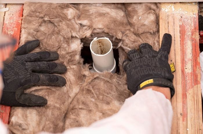 A person wearing gloves is fitting insulation around a pipe by cutting a hole in the material to ensure a snug fit. 