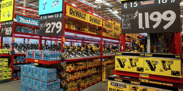 Power tool section of a hardware warehouse with prominent DEWALT, Makita, Ryobi and Ozito displays. Red shelving is filled with boxed tools and accessories, while large overhead signs highlight product ranges and promotional pricing.