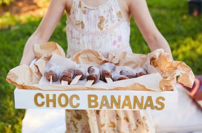 Child holding a painted wooden crate tray labelled ‘choc bananas’ with chocolate-coated frozen bananas on display