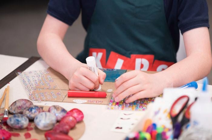 A kid is applying glue to a coaster board with craft rhinestone stickers and alphabets on the table.