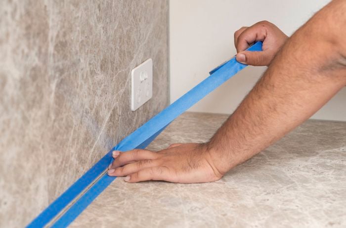 Hands remove blue masking tape from the joint between a stone-look benchtop and splashback, revealing a neat silicone seal along the edge.