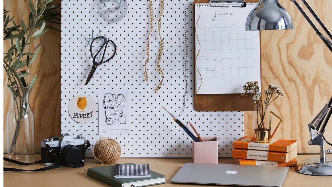 An eclectic workspace with a pegboard against the far wall, showing books, a laptop, a camera, a ball of twine, a clipboard calendar, scissors, a Pixar desk lamp, a tree branch in a vase, a plastic chakram ruler and strips of lace