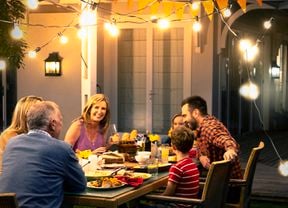  A group of people seated around an outdoor dining table on a verandah at night eating dinner with festoon lights above and a Jackery Power Station in the background.