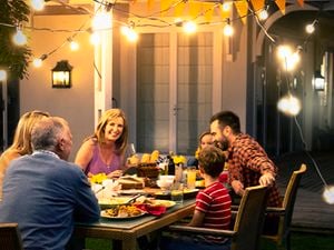  A group of people seated around an outdoor dining table on a verandah at night eating dinner with festoon lights above and a Jackery Power Station in the background.