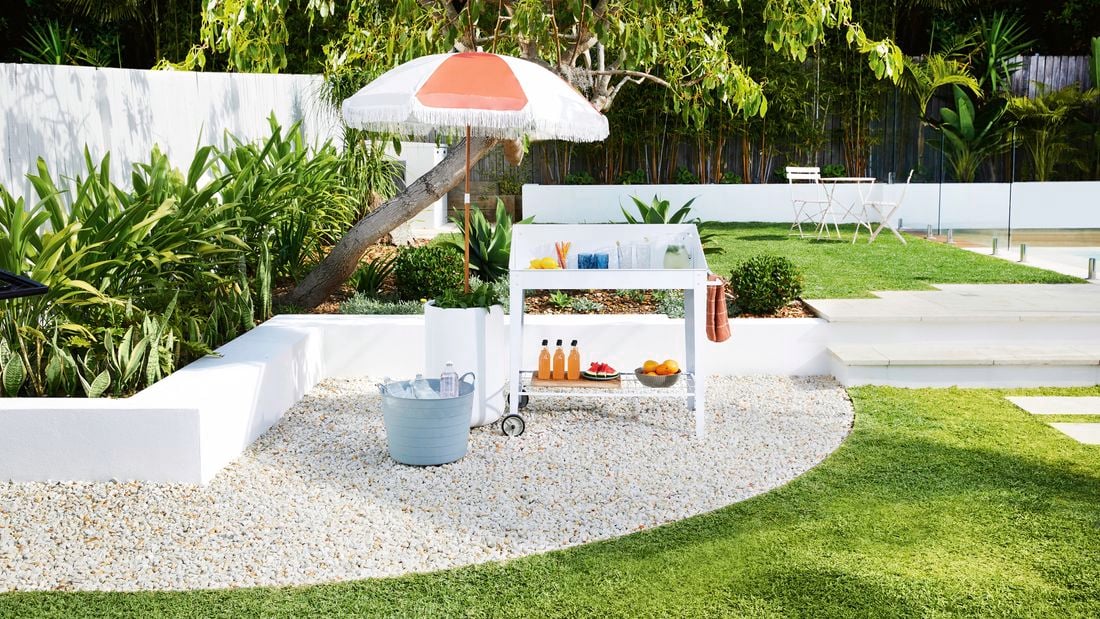A white drink trolley sits in a backyard, set up with fruit, glasses and drinks, next to a red and white shade umbrella.