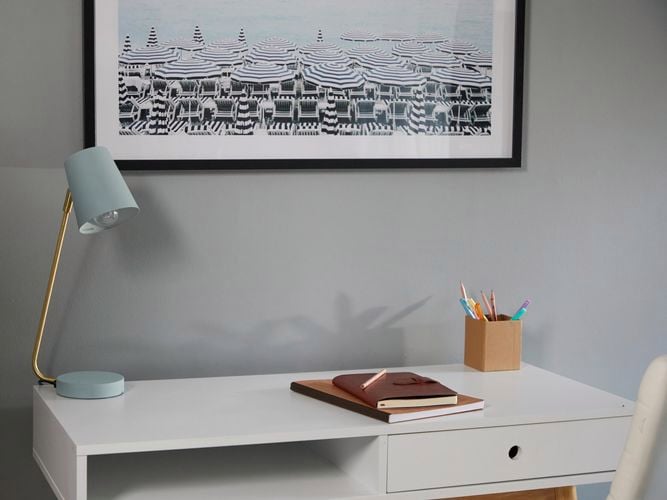Painted metal desk lamp sits on a desk in front of a grey wall.