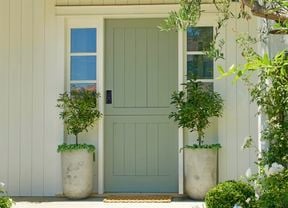 A light green front door with a black smart lock, flanked by two potted trees, and surrounded by white siding and garden foliage.