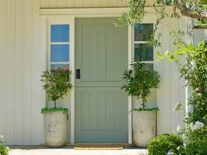 A light green front door with a black smart lock, flanked by two potted trees, and surrounded by white siding and garden foliage.