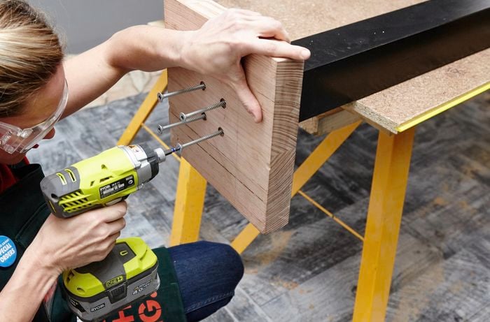 Four long screws being drilled into the base of a wooden coat rack by a Bunnings team member with a power drill