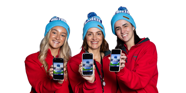 Three people wearing red jackets and blue beanies stand against a plain background, each holding up a smartphone displaying a “Big Freeze” fundraising page.