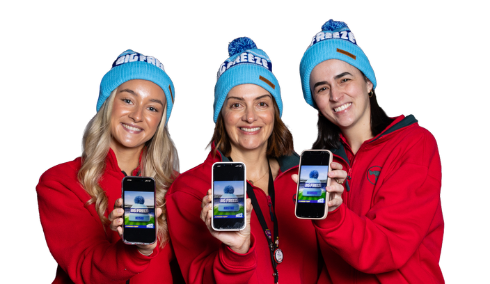 Three people wearing red jackets and blue beanies stand against a plain background, each holding up a smartphone displaying a “Big Freeze” fundraising page.