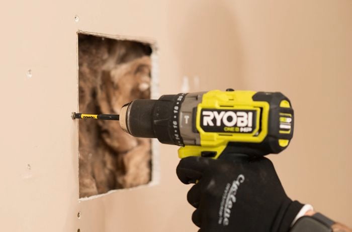 A gloved hand is using a Ryobi screw gun to install screws into the plasterboard.
