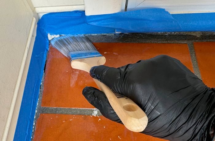 A hand wearing a glove is using a paintbrush to cut in around the edges of the tiles with primer