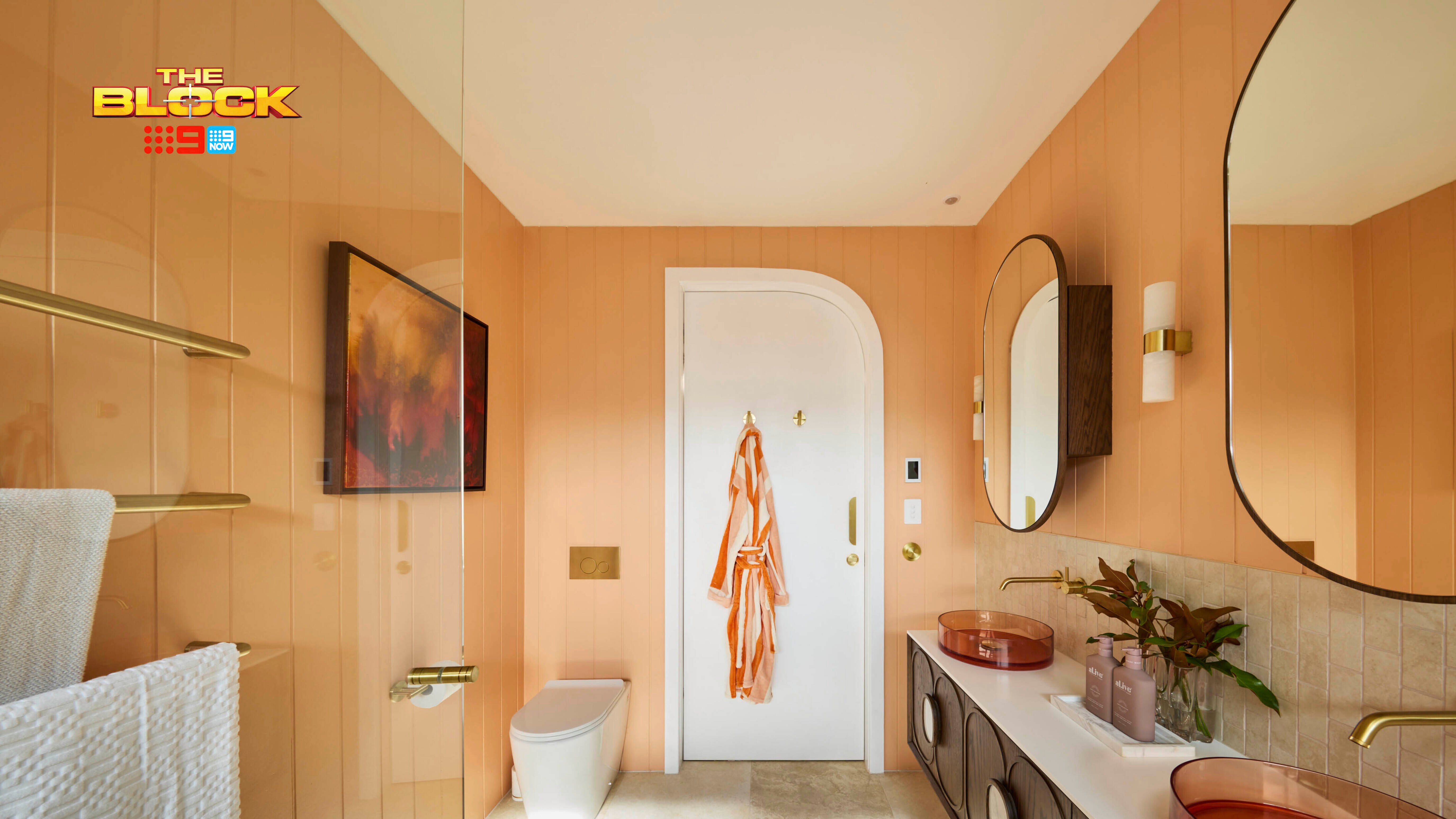 Modern bathroom with peach walls, double copper sinks, round mirrors, gold fixtures, white toilet, and orange robe on door.