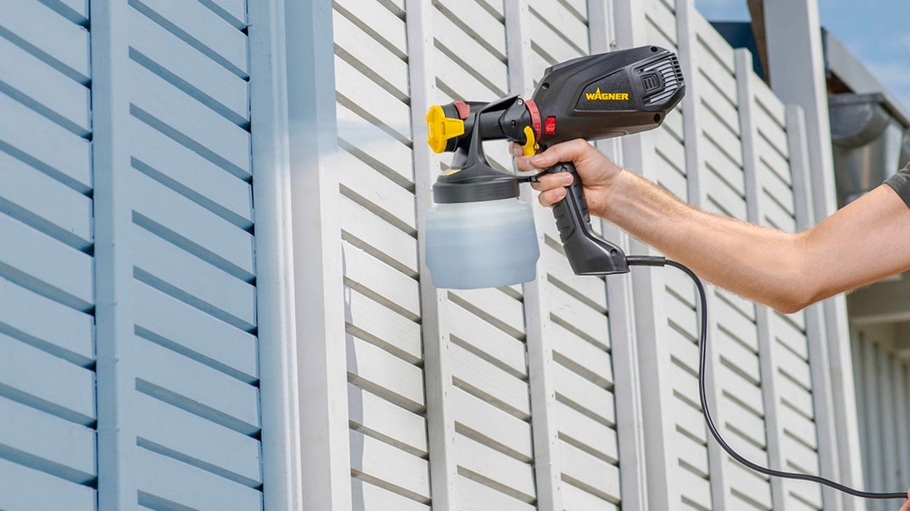  A pair of hands holding a nail and staple gun onto a white fence in front of a green bush.