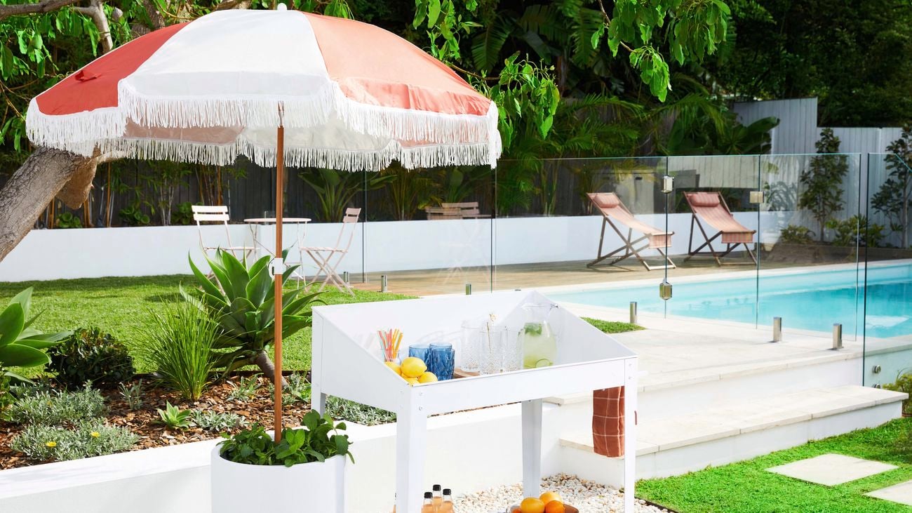 A white drinks trolley sits in a backyard with a pull, set up with fruit and glasses on its' top and bottom levels, next to a red and white shade umbrella.