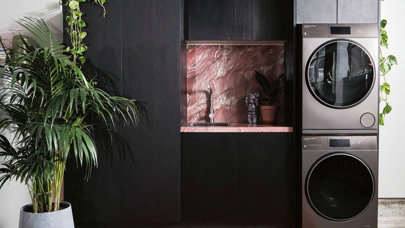 A laundry room with stacked appliances, a sink, dark cabinetry, plants, and an ornate rug.